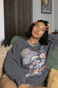 Model Aina sits on a couch in a grey sweatshirt, has green eyeshadow and multicolored butterfly hair clips.