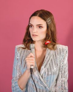 Jane Ellen Bryant Stand in front of Pink backdrop with a sequined jacket on while holding a microphone.